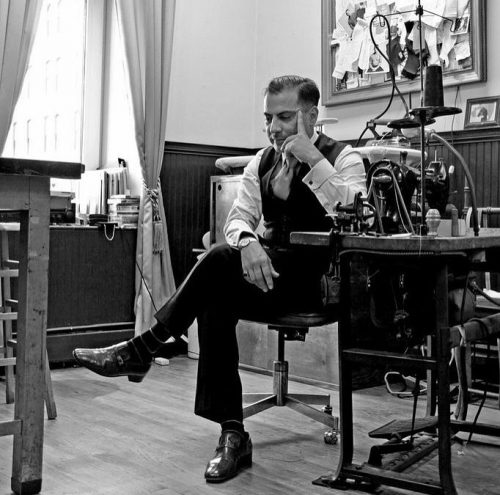 Man dressed in a suit with his legs crossed in a tailor shop. He is seated in a pensive pose next to a sewing machine
